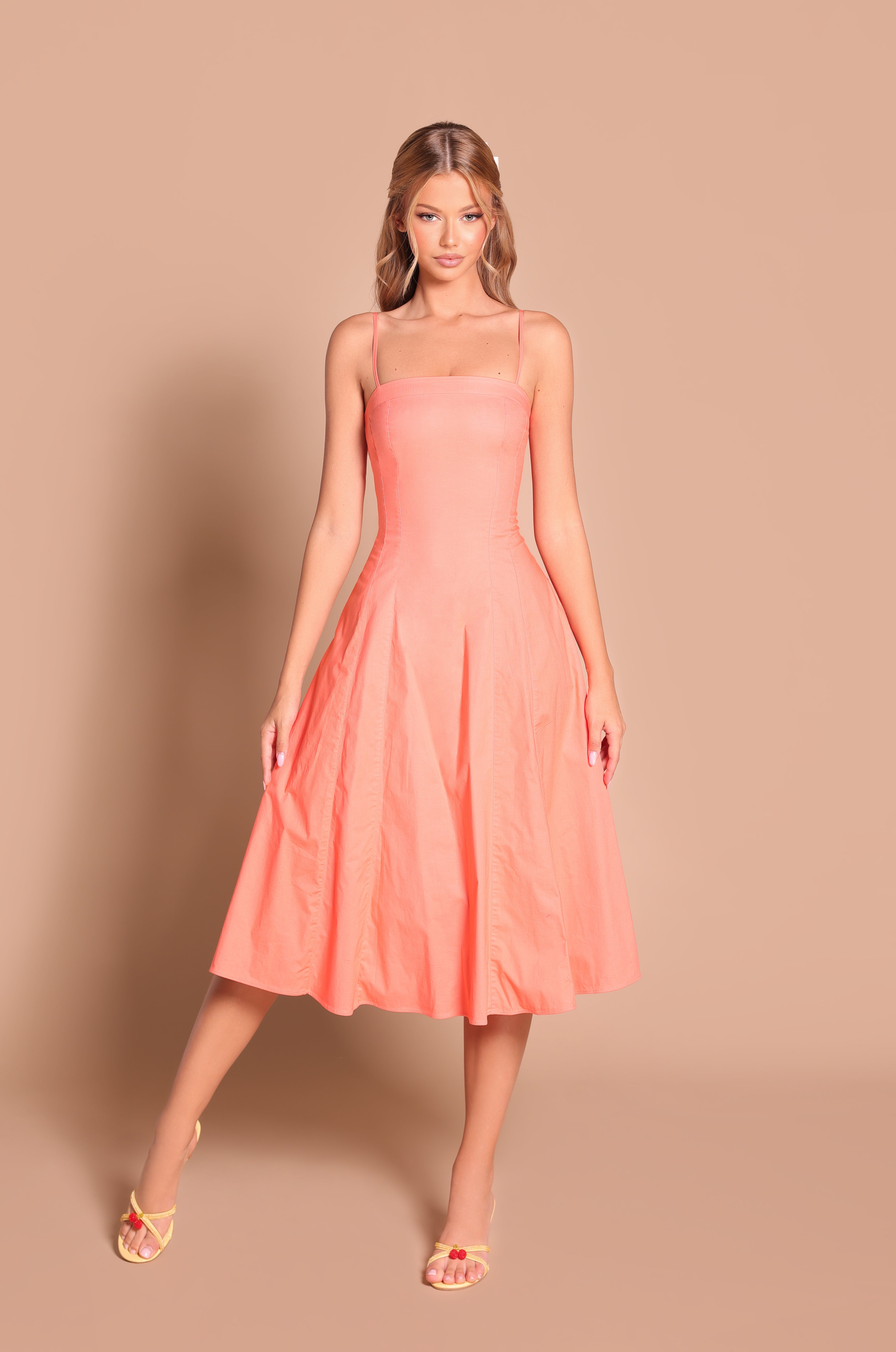 A woman stands against a beige background, wearing the Papaya Midi Casual Summer Dress: Auberine, a sleeveless peach midi dress paired with light yellow open-toe heels.